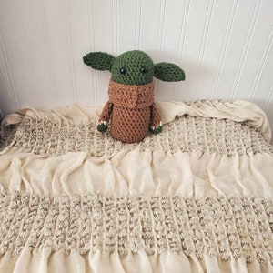 May include: A crocheted green and brown Baby Yoda plush toy sitting on a white crocheted blanket.