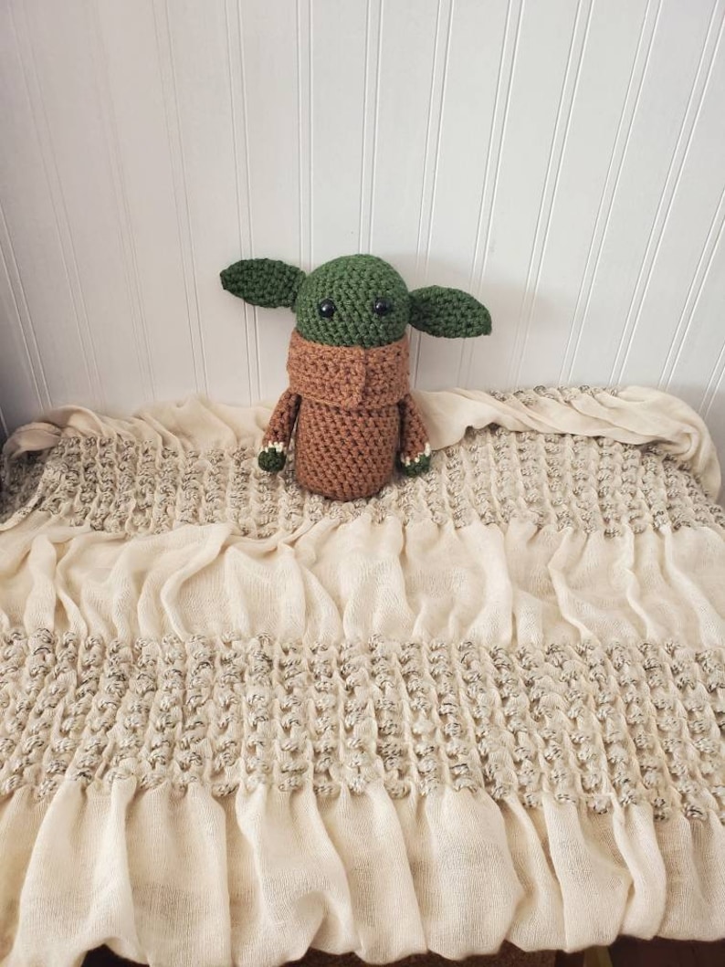 May include: A crocheted green and brown Baby Yoda plush toy sitting on a white crocheted blanket.