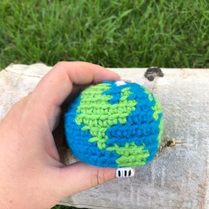 May include: A crocheted globe with blue and green yarn, representing the Earth. The globe is held in a hand with a white nail with black piano keys painted on it.