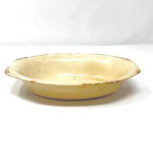 May include: A vintage, oval-shaped ceramic baking dish with a light yellow glaze. The dish has a slightly raised rim and shows signs of age with brown discoloration around the edges. The interior is smooth and the overall design is simple and functional.