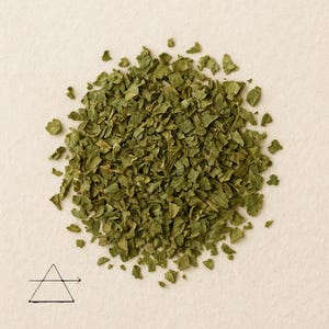 May include: A pile of dried, chopped green parsley flakes. The flakes are a medium green color and have a slightly textured appearance. A black and white triangle with a line and arrow is in the lower left corner.