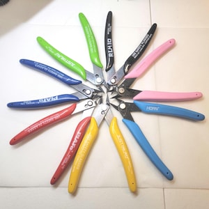 May include: A selection of angled wire cutters with various coloured handles, including green, blue, pink, yellow, and red. Each cutter has a silver blade and text that reads "USE SAFETY GLASSES" and brand names like "PLATO" and "JUSTINEAU".