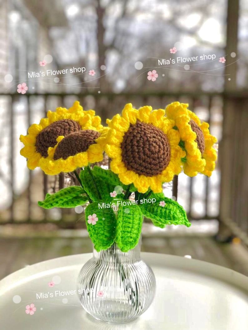 Peut inclure: Un bouquet de tournesols en crochet dans un vase en verre transparent &agrave; texture c&ocirc;tel&eacute;e. Les tournesols sont jaunes avec des centres bruns et des feuilles vertes. Le vase est pos&eacute; sur une table blanche. Le texte "Mia's Flower shop" appara&icirc;t plusieurs fois dans l'image.