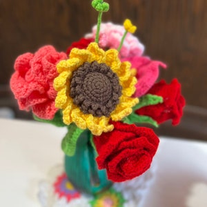 Peut inclure: Un bouquet de fleurs crochet&eacute;es dans un vase vert. Le bouquet comprend un tournesol, des fleurs roses et des roses rouges.