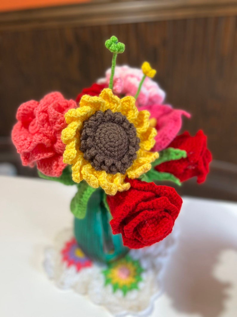 Peut inclure: Un bouquet de fleurs crochet&eacute;es dans un vase vert. Le bouquet comprend un tournesol, des fleurs roses et des roses rouges.