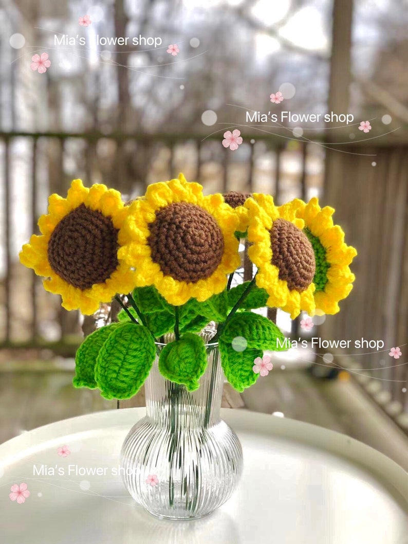Peut inclure: Un bouquet de tournesols en crochet dans un vase en verre transparent avec une surface textur&eacute;e. Les tournesols sont jaunes avec des centres bruns et des feuilles vertes. Le vase est pos&eacute; sur une table blanche.