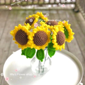 Peut inclure: Un bouquet de tournesols en crochet dans un vase en verre transparent. Les tournesols sont jaunes avec des centres bruns et des feuilles vertes. Le vase est pos&eacute; sur une table blanche. Le texte "Mia's Flower shop" est &eacute;crit sur l'image.