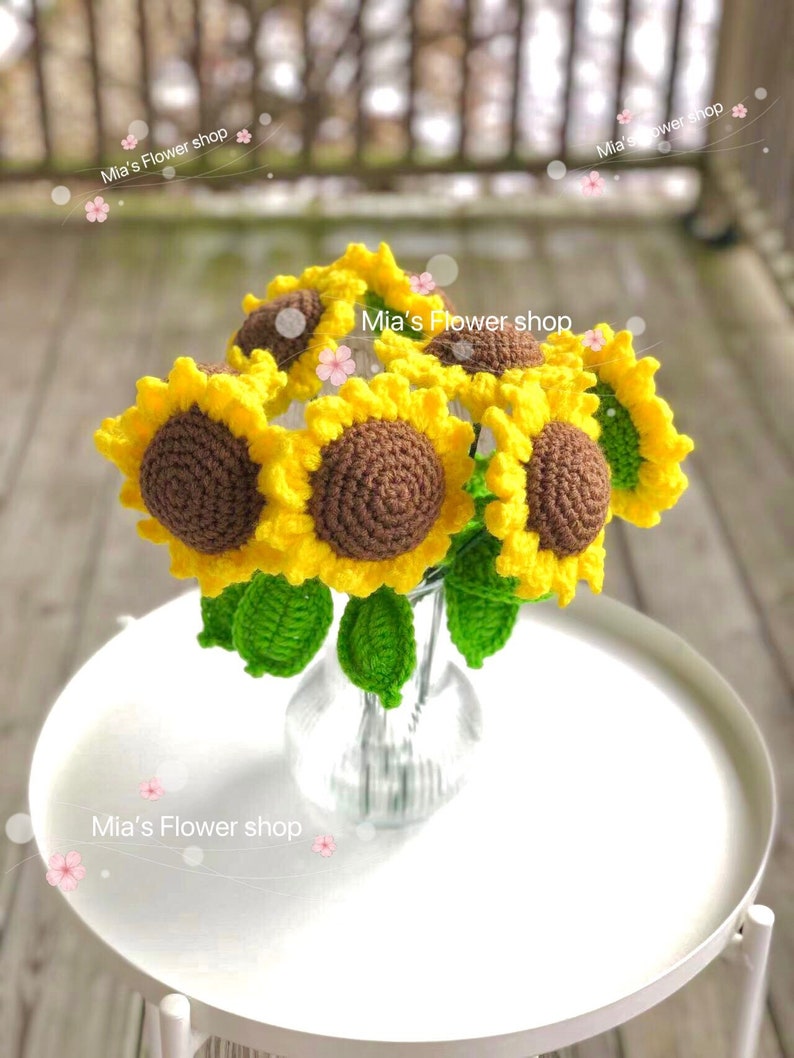 Peut inclure: Un bouquet de tournesols en crochet dans un vase en verre transparent. Les tournesols sont jaunes avec des centres bruns et des feuilles vertes. Le vase est pos&eacute; sur une table blanche. Le texte "Mia's Flower shop" est &eacute;crit sur l'image.