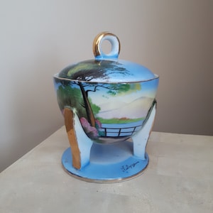 Peut inclure: Un sucrier en porcelaine bleu et blanc avec un couvercle et un pied dorés. Le bol est décoré d'une scène de paysage peinte représentant un pont, des arbres et des montagnes. Le fond du bol est signé "J. Lopez".