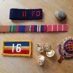 Op de afbeelding: Een verzameling militaire patches en badges, waaronder een rood-wit-blauwe patch met het nummer "16" erop, een zwarte patch met de tekst "II FD", een rood-wit-blauw lint, een gouden metalen badge met de tekst "LOGISTICS SERVITIUM NULLI SECUNDUS LOGISTIQUE", en vier gouden knopen.
