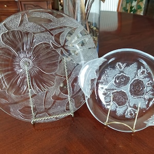 May include: Two clear glass serving dishes with floral designs. The larger dish features a large flower with leaves, while the smaller dish has a cluster of roses with leaves. Both dishes are on decorative stands.