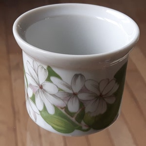 May include: White ceramic cup with a floral design featuring white flowers with pink centers and green leaves.