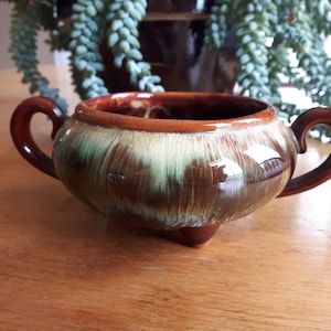 May include: A ceramic sugar bowl in shades of brown and green, featuring two handles and three small feet. The bowl has a glossy finish and a brown rim. The exterior has a mottled green and brown pattern. The bowl is likely for serving sugar.