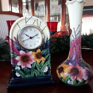 May include: A floral clock and vase with a white background and colourful flowers. The clock has a gold rim and black hands. The vase is a light cream colour with a ruffled top.