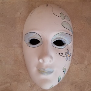 May include: White ceramic mask with a painted design of a flower and leaves. The mask has glitter accents around the eyes and mouth.