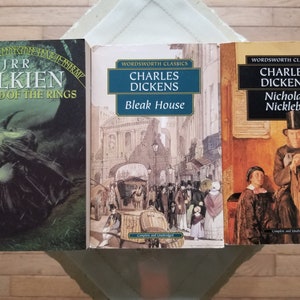 May include: Three paperback books on a wooden surface. The book on the left is "The Lord of the Rings" by J.R.R. Tolkien. The middle book is "Bleak House" by Charles Dickens. The book on the right is "Nicholas Nickleby" by Charles Dickens.