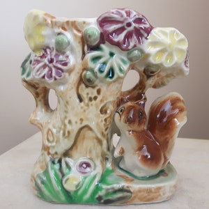 May include: A ceramic vase shaped like a tree trunk with a squirrel sitting at the base. The vase is decorated with flowers in shades of red, yellow, and green.