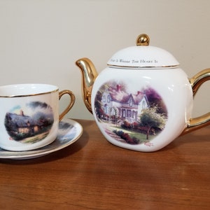 May include: A white tea set with gold accents featuring a cottage scene. The teapot has the text "Home Is Where The Heart Is" on the lid. The cup and saucer have a different scene of a cottage in the woods.