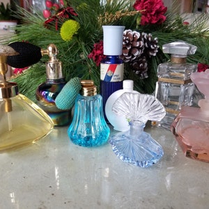 May include: A collection of vintage perfume bottles in various colors and styles, including clear glass, blue glass, and pink glass. Some bottles have decorative stoppers and atomizers. The bottles are arranged on a table with a green garland in the background.