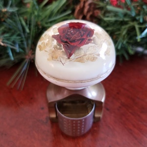 Könnte beinhalten: Ein weißer Keramik-Knopf mit goldener Einfassung und einer roten Rose, die in klarem Harz eingebettet ist. Der Knopf ist an einer Messingbasis mit einem kleinen, silberfarbenen Becher befestigt.