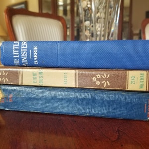 May include: A stack of three vintage hardback books with blue and brown covers. The top book is titled "The Little Minister" by Barrie. The middle book is titled "Eight Cousins" by Alcott. The bottom book has the number "2143" and the name "Whitman" printed on the spine.