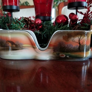 Może przedstawiać: Biała porcelana z dwoma uchwytami, przedstawiająca malowaną scenę krajobrazową z domem nad jeziorem o zachodzie słońca. Naczynie ma czarną obwódkę i jest ozdobione delikatnym wzorem kwiatowym.