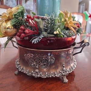 May include: A silver and glass centrepiece with a red glass bowl and a silver base with ornate detailing. The bowl is filled with greenery, red berries, and gold ornaments.