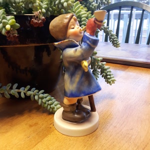 May include: A porcelain figurine of a child wearing a blue coat and a red hat, holding a horn and a stick. The figurine is standing on a white base.