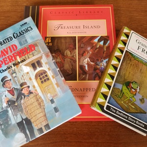 May include: Three hardback books on a wooden surface. The book on the left is blue and features a picture of two men in Victorian era clothing. The title is "David Copperfield" by Charles Dickens. The book in the middle is red and features a picture of a pirate ship. The title is "Treasure Island" by R.L. Stevenson. The book on the right is green and features a picture of a frog. The title is "The Adventures of Grandfather Frog" by Thornton W. Burgess.