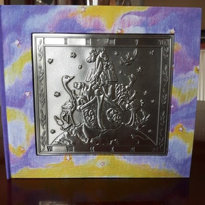 May include: A purple and yellow book cover with a silver embossed image of Noah's Ark with animals on top. The ark is surrounded by a silver frame with Roman numerals.