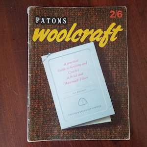 May include: A blue and white book titled "Patons Woolcraft" with the text "A practical Guide to Knitting and Crochet in Wool and Man-made Fibres" and "18th Edition" on the cover. The book is on a brown wooden surface.