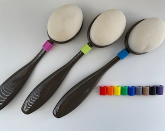 Puede incluir: Tres pinceles de maquillaje marrones con puntas de esponja ovaladas blancas. Los pinceles tienen anillos de colores diferentes alrededor de los mangos: rosa, verde y azul. Una fila de diez anillos de plástico de colores se encuentra debajo de los pinceles.