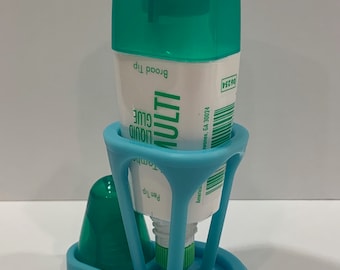 May include: A blue plastic dispenser for a tube of liquid glue. The dispenser has a base with the words "YOUR MESSAGE" printed on it. The glue tube is labelled "Tombe MULTI LIQUID GLUE" and has a green cap.