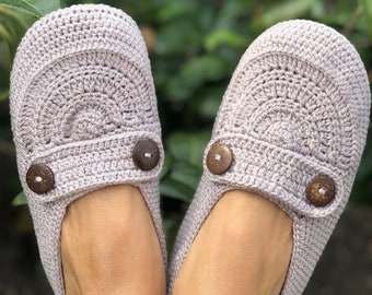 May include: A pair of light gray crocheted slippers with brown button accents. The slippers are worn on a person's feet.