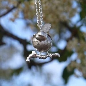 Könnte beinhalten: Eine silberne Halskette mit einem kleinen Vogel-Anhänger. Der Vogel sitzt auf einem Ast und hat ein detailliertes Design. Die Halskette hängt an einem Ast mit weißen Blüten.