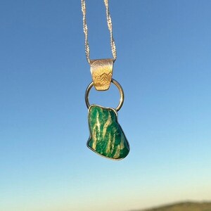 May include: A silver necklace with a green and white stone pendant. The pendant is an irregular shape, suspended from a silver ring. The necklace hangs against a backdrop of a green field and a blue sky.