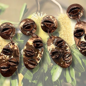 Op de afbeelding: Vier paar koperen oorbellen met een uniek ontwerp met meerdere afgeronde vormen. De oorbellen worden getoond tegen een achtergrond van gedroogde bloemen.