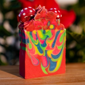 May include: A vibrant, handmade soap bar with a red base and colorful swirls of blue, yellow, and green. The top is decorated with red mushroom-shaped accents with white polka dots. A unique, decorative item.