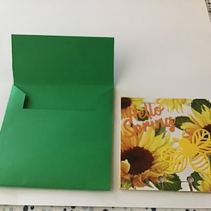 May include: A green card with a white card inside. The white card has a sunflower pattern and the text "Hello Spring" in orange. A yellow butterfly cutout is on the card.