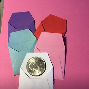 May include: Five small origami envelopes in different colors, including purple, red, pink, blue, and white. A silver US quarter is placed on top of the white envelope.
