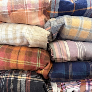 Puede incluir: Una pila de camisas de franela dobladas en varios patrones de cuadros. Los colores incluyen naranja, rojo, azul y beige. Las camisas están cuidadosamente apiladas, mostrando las diferentes combinaciones de colores y texturas de la tela.