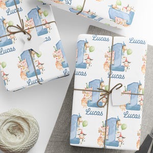 May include: White wrapping paper featuring a blue number one and the name "Lucas". The paper is decorated with cartoon animals and birthday party elements. The wrapped gifts are tied with twine and a blank tag.