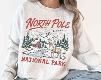 May include: An ash gray crewneck sweatshirt featuring a graphic print with the text North Pole National Park in red. The illustration depicts a snowy mountain landscape, a small red cabin, evergreen trees, and a reindeer standing in the foreground.
