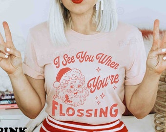 May include: A light pink t-shirt with the text "I See You When You're Flossing" and a Santa Claus graphic in red. The model is wearing a red and white striped headband and shorts.