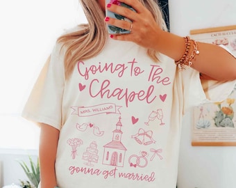 Könnte beinhalten: Ein elfenbeinfarbenes T-Shirt mit rosa Schriftzug, der "Going to the Chapel" und "gonna get married" lautet. Das Shirt zeigt außerdem eine rosa Illustration einer Kirche, Eheringe, Vögel, Kuchen und andere Hochzeitsmotive.