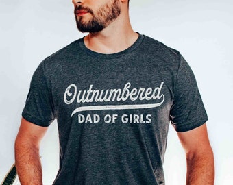 May include: A dark grey heather t-shirt with the text "Outnumbered Dad of Girls" in white.