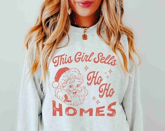 Puede incluir: Sudadera gris claro con un gráfico rojo que dice "This Girl Sells Ho Ho Homes" con la cara de Santa Claus. La sudadera es de cuello redondo.