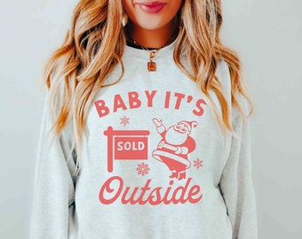 Puede incluir: Sudadera gris claro con un gráfico rojo que dice "BABY IT'S SOLD Outside". El gráfico presenta una figura de Papá Noel, copos de nieve y un letrero de "SOLD". La palabra "ash" está impresa en la esquina inferior izquierda.
