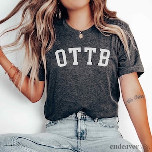 May include: Dark gray t-shirt with the white letters OTTB across the chest. The shirt is a crew neck style and appears to be made of a soft material. The shirt is paired with blue jeans.
