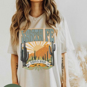 May include: A light gray t-shirt with a graphic print of a sunset scene with cacti and the text "SANTA FE" and "SARA'S BACH" in a retro font.